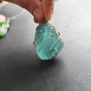 May include: A teal-colored, irregularly shaped pendant, wrapped in silver-colored wire. The pendant is held up by a hand, and the background is a dark surface. The pendant appears to be made of sea glass.