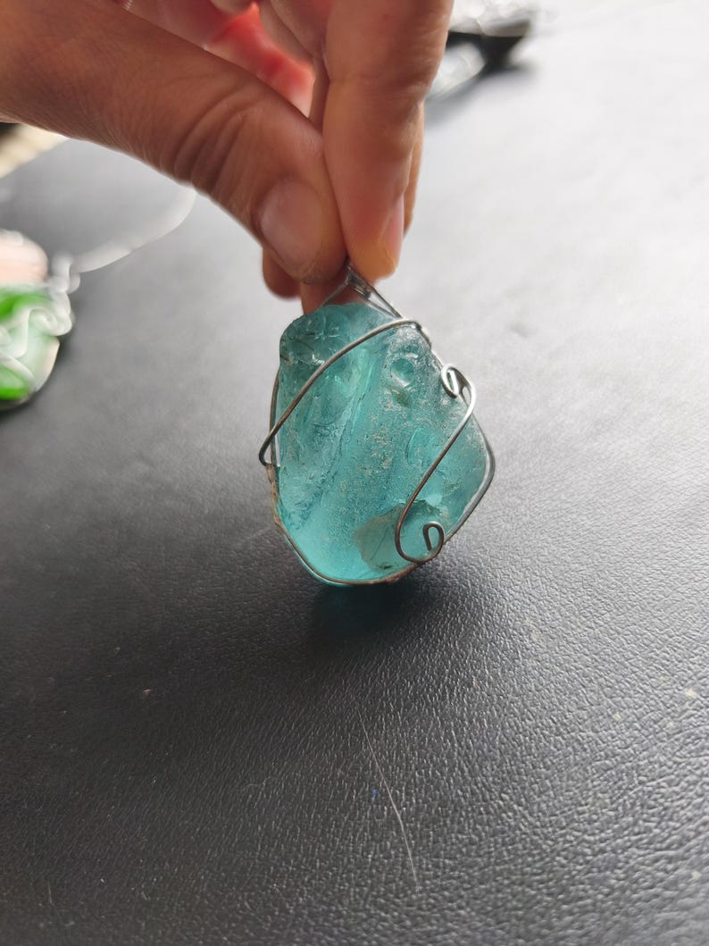 May include: A teal-colored, irregularly shaped pendant, wrapped in silver-colored wire. The pendant is held up by a hand, and the background is a dark surface. The pendant appears to be made of sea glass.