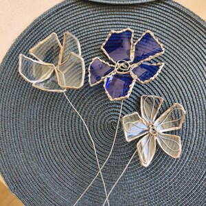 May include: Three decorative glass flowers with silver wire stems. One flower is a deep blue, the other two are clear. The flowers are arranged on a dark blue textured placemat. The glass petals are outlined in silver.