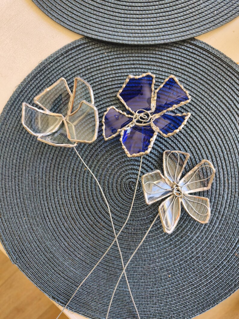 May include: Three decorative glass flowers with silver wire stems. One flower is a deep blue, the other two are clear. The flowers are arranged on a dark blue textured placemat. The glass petals are outlined in silver.