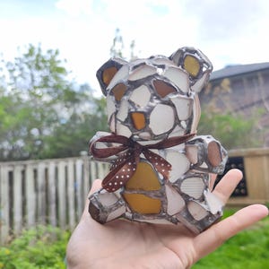 May include: A handcrafted teddy bear figurine made from sea glass pieces in shades of white, amber, and brown, held in a hand. The bear has a brown ribbon with white polka dots tied around its neck. The sea glass is set in a silver-colored metal.