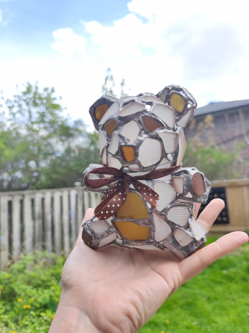 May include: A handcrafted teddy bear figurine made from sea glass pieces in shades of white, amber, and brown, held in a hand. The bear has a brown ribbon with white polka dots tied around its neck. The sea glass is set in a silver-colored metal.