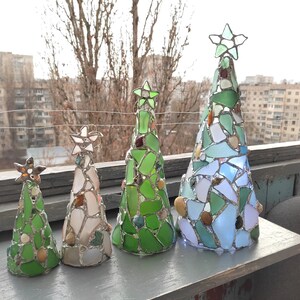 Pode incluir: Quatro esculturas decorativas de &aacute;rvores de Natal. Cada &aacute;rvore tem formato c&ocirc;nico e &eacute; feita de pe&ccedil;as de vidro coloridas, com uma estrela no topo. As &aacute;rvores variam em tamanho e apresentam tons de verde, branco e azul, com pequenas pedras.