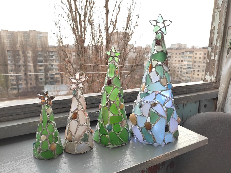 Pode incluir: Quatro esculturas decorativas de &aacute;rvores de Natal. Cada &aacute;rvore tem formato c&ocirc;nico e &eacute; feita de pe&ccedil;as de vidro coloridas, com uma estrela no topo. As &aacute;rvores variam em tamanho e apresentam tons de verde, branco e azul, com pequenas pedras.