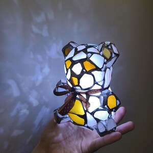 May include: A mosaic teddy bear lamp with a brown ribbon around its middle. The lamp is made of white, yellow, and orange glass pieces. The lamp is lit from within, creating a warm glow.