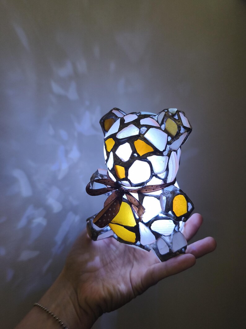 May include: A mosaic teddy bear lamp with a brown ribbon around its middle. The lamp is made of white, yellow, and orange glass pieces. The lamp is lit from within, creating a warm glow.