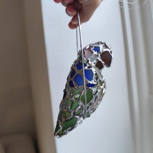 May include: A stained glass parrot ornament with a silver metal frame. The bird features blue, green, and brown glass pieces. The ornament is suspended by a silver wire loop, suitable for hanging. The parrot's design is a mosaic of colorful glass.