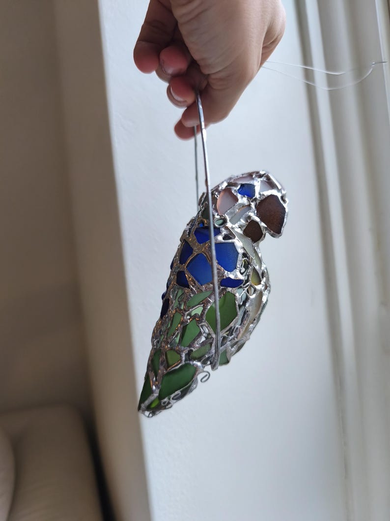 May include: A stained glass parrot ornament with a silver metal frame. The bird features blue, green, and brown glass pieces. The ornament is suspended by a silver wire loop, suitable for hanging. The parrot's design is a mosaic of colorful glass.