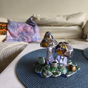 May include: A decorative mushroom-shaped lamp with a stained glass design. The mushroom caps are brown and amber, and the base features green glass accents. The lamp sits on a blue placemat, with a sofa and pillows in the background.