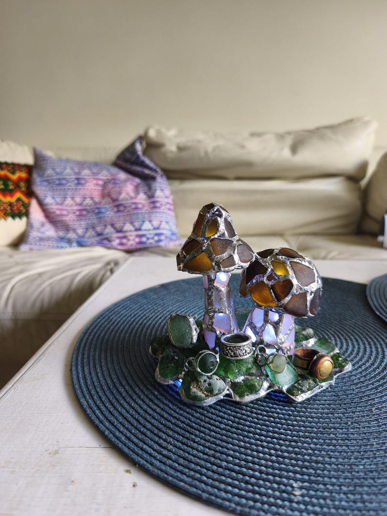 May include: A decorative mushroom-shaped lamp with a stained glass design. The mushroom caps are brown and amber, and the base features green glass accents. The lamp sits on a blue placemat, with a sofa and pillows in the background.