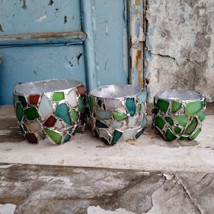 Könnte beinhalten: Drei dekorative Pflanzgefäße mit Mosaikdesign. Die Pflanzgefäße bestehen aus Glasstücken in Grün-, Braun- und Weißtönen, die in einem silberfarbenen Metallrahmen gefasst sind. Der größte Topf hat einen Durchmesser von etwa 15 cm.