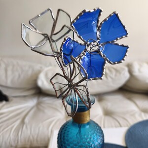 May include: A decorative glass flower sculpture featuring blue and clear petals, mounted on a turquoise glass bottle. The petals are edged with silver solder. The bottle has a gold band around the neck. The sculpture is a unique floral arrangement.