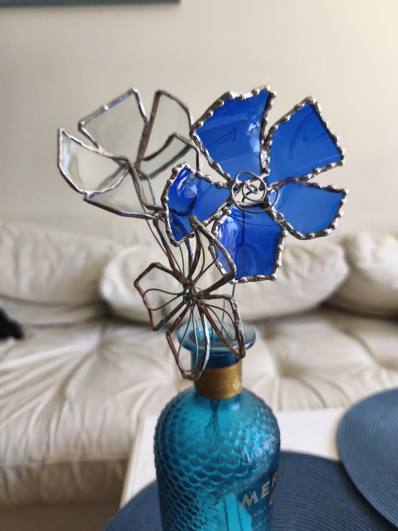 May include: A decorative glass flower sculpture featuring blue and clear petals, mounted on a turquoise glass bottle. The petals are edged with silver solder. The bottle has a gold band around the neck. The sculpture is a unique floral arrangement.