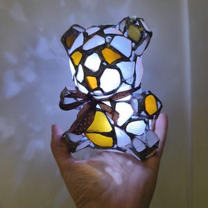 May include: A mosaic-style teddy bear lamp with a brown ribbon. The lamp is made of white, yellow, and brown glass pieces. The lamp is lit from within, creating a warm glow.