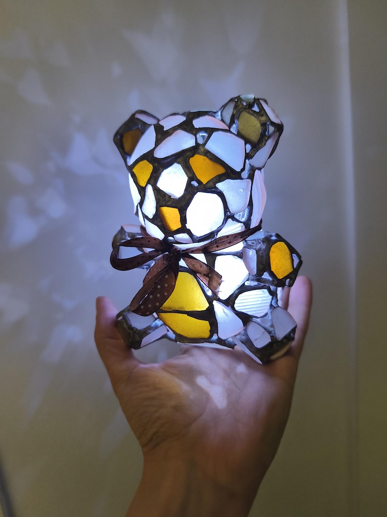 May include: A mosaic-style teddy bear lamp with a brown ribbon. The lamp is made of white, yellow, and brown glass pieces. The lamp is lit from within, creating a warm glow.
