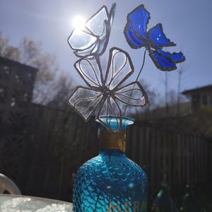 May include: A blue glass bottle, labelled "MERMAID GIN", supports a glass flower arrangement. The flowers and butterfly are clear and blue, with dark metal stems. The arrangement is set against a bright sunny sky, with the sun shining through the glass.