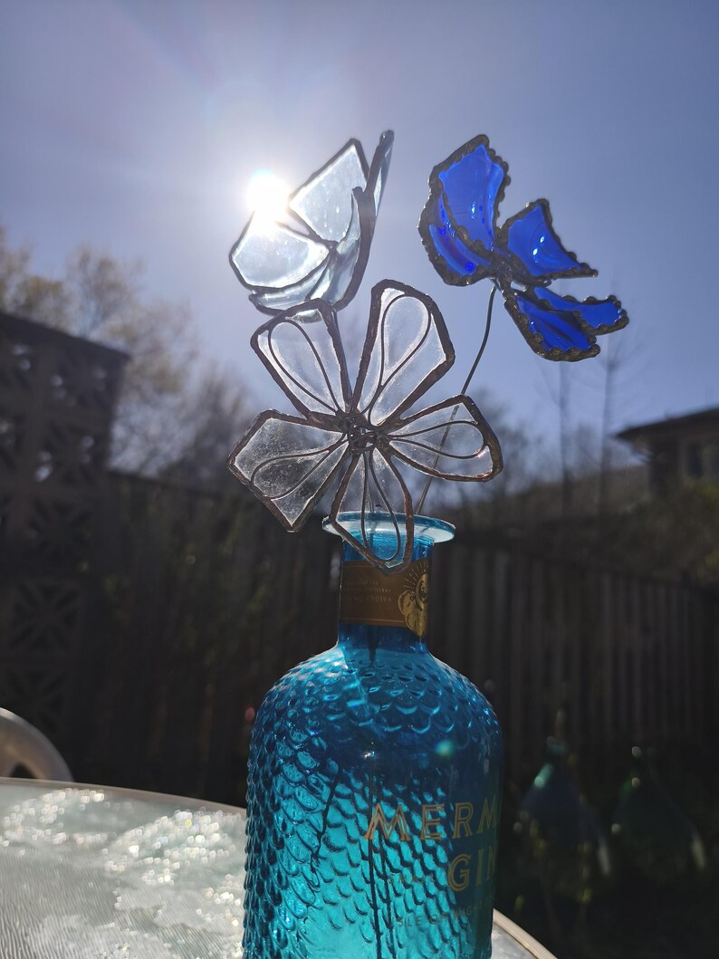 May include: A blue glass bottle, labelled "MERMAID GIN", supports a glass flower arrangement. The flowers and butterfly are clear and blue, with dark metal stems. The arrangement is set against a bright sunny sky, with the sun shining through the glass.