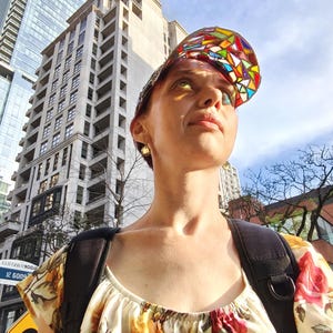 May include: A woman wearing a colorful stained-glass visor cap and a floral print top, with a black backpack. The background features tall buildings and a sunny sky, suggesting an urban setting.