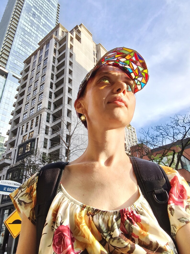 May include: A woman wearing a colorful stained-glass visor cap and a floral print top, with a black backpack. The background features tall buildings and a sunny sky, suggesting an urban setting.