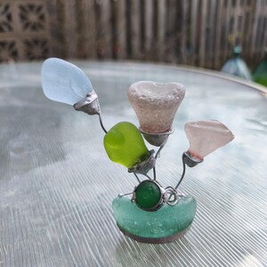 May include: A decorative sculpture made from sea glass in blue, green, and pink hues. The sea glass is held together with silver-coloured wire, creating a floral design. A green, round stone is set in the centre of the base.