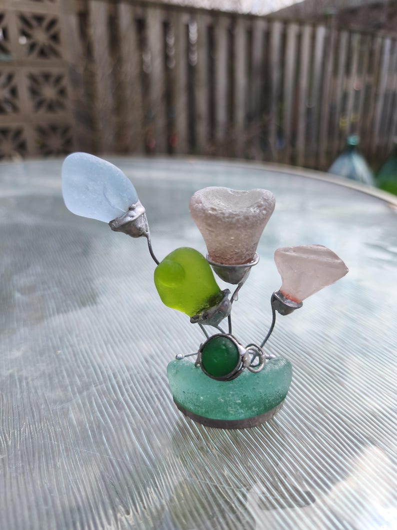 May include: A decorative sculpture made from sea glass in blue, green, and pink hues. The sea glass is held together with silver-coloured wire, creating a floral design. A green, round stone is set in the centre of the base.