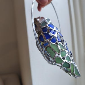 May include: A handcrafted, decorative piece featuring a mosaic design of blue and green glass pieces set in a silver-colored metal frame. The glass pieces are irregular in shape and size, creating a unique, textured appearance. The piece is held by a silver metal hoop.