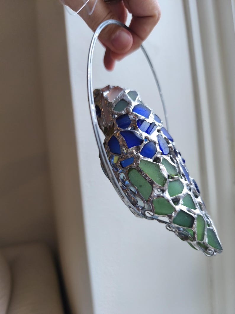May include: A handcrafted, decorative piece featuring a mosaic design of blue and green glass pieces set in a silver-colored metal frame. The glass pieces are irregular in shape and size, creating a unique, textured appearance. The piece is held by a silver metal hoop.