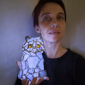May include: A mosaic cat figurine with a gold trim and yellow eyes. The cat is made of white and blue glass pieces, and it is illuminated from within. The figurine is held in a person's hand.