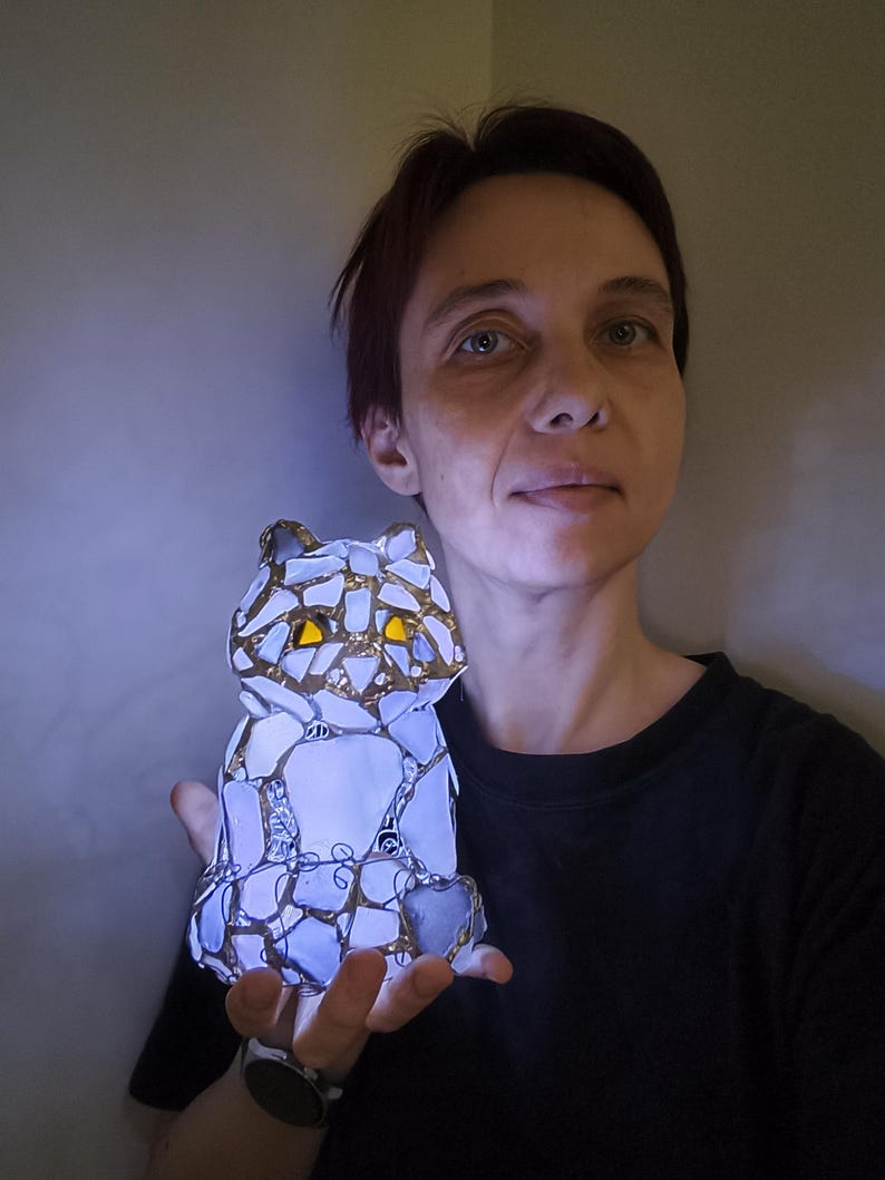 May include: A mosaic cat figurine with a gold trim and yellow eyes. The cat is made of white and blue glass pieces, and it is illuminated from within. The figurine is held in a person's hand.