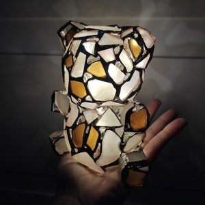 May include: A mosaic teddy bear figurine with a patchwork design of white, gold, and black pieces. The bear is illuminated from within, creating a soft glow. The bear is held in a hand.