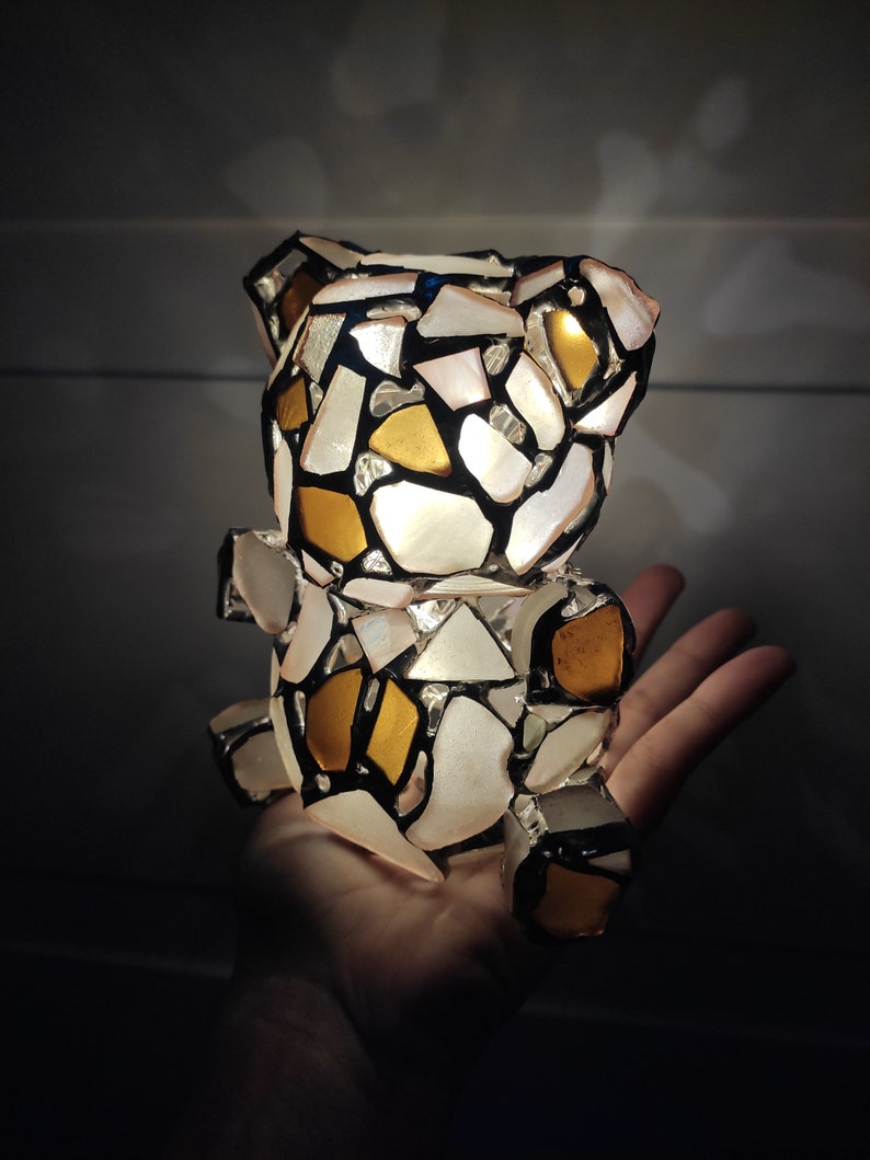 May include: A mosaic teddy bear figurine with a patchwork design of white, gold, and black pieces. The bear is illuminated from within, creating a soft glow. The bear is held in a hand.