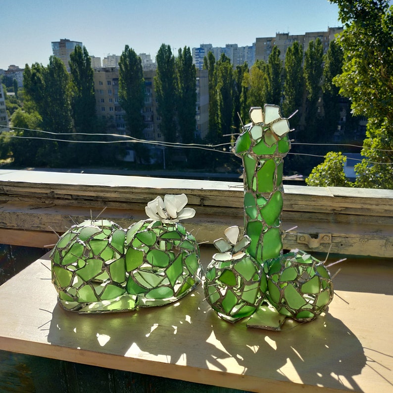 May include: A green glass sculpture of a cactus with white glass flowers. The sculpture is made of many small pieces of glass that are joined together.