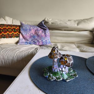 May include: A decorative glass mushroom sculpture with brown and iridescent caps and green base. The sculpture sits on a blue placemat on a white table. A white couch and patterned pillows are in the background.