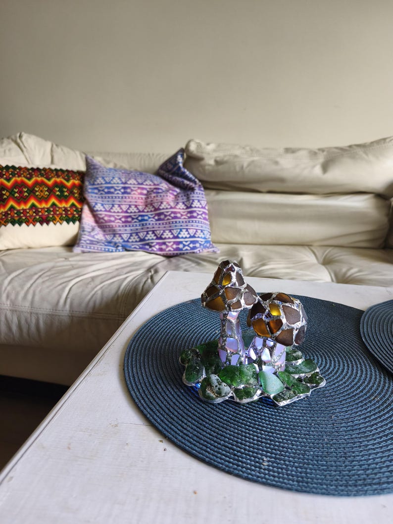 May include: A decorative glass mushroom sculpture with brown and iridescent caps and green base. The sculpture sits on a blue placemat on a white table. A white couch and patterned pillows are in the background.