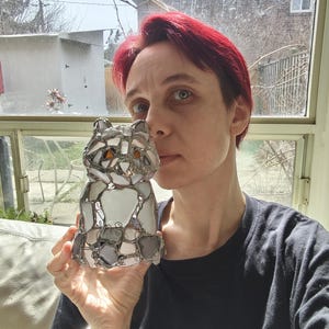 May include: A mosaic cat sculpture with a silver metal frame. The cat is made of various shapes of white, grey, and orange glass pieces. The sculpture is a decorative item, possibly for a home or garden.