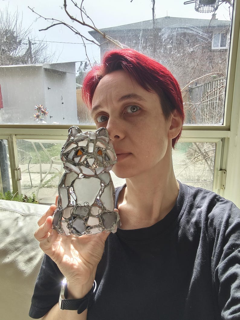 May include: A mosaic cat sculpture with a silver metal frame. The cat is made of various shapes of white, grey, and orange glass pieces. The sculpture is a decorative item, possibly for a home or garden.