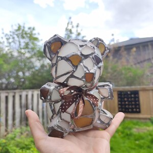 May include: A handcrafted bear figurine made of sea glass pieces in white, brown, and amber hues, with a brown polka-dot ribbon tied around its neck. The bear is held in a hand against a backdrop of greenery and a cloudy sky.