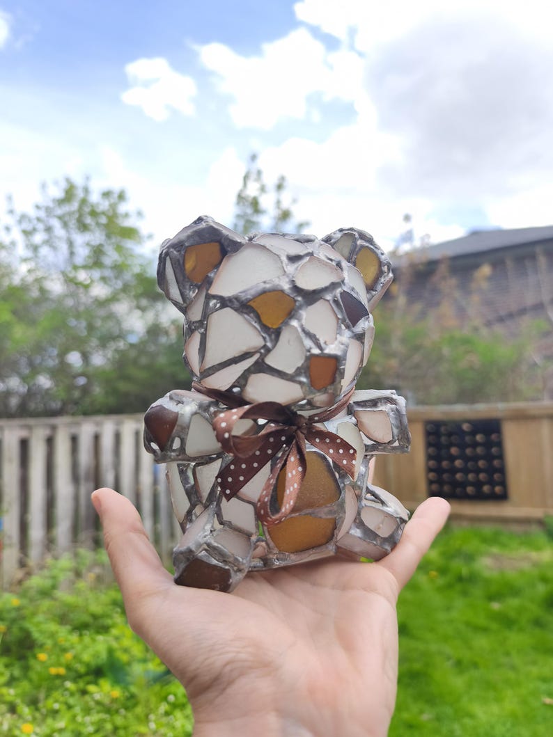 May include: A handcrafted bear figurine made of sea glass pieces in white, brown, and amber hues, with a brown polka-dot ribbon tied around its neck. The bear is held in a hand against a backdrop of greenery and a cloudy sky.