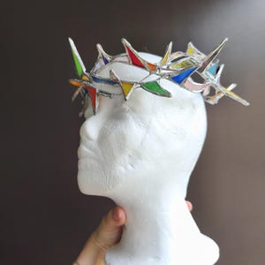 May include: A crown constructed from multiple triangular pieces of multicolored stained glass held together by silver metal solder. The crown features sharp, pointed edges and a circular band design, showcasing various hues including red, blue, green, and yellow.