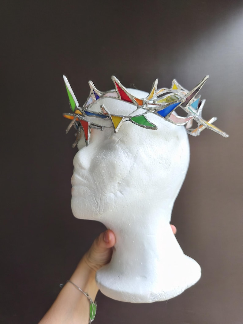 May include: A crown constructed from multiple triangular pieces of multicolored stained glass held together by silver metal solder. The crown features sharp, pointed edges and a circular band design, showcasing various hues including red, blue, green, and yellow.