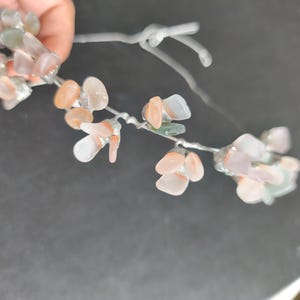 May include: A delicate floral hair accessory featuring small, translucent, pastel-colored stones. The stones are in shades of peach, pink, and green, attached to a silver-colored wire, creating a natural, organic look. The accessory is held by a hand.
