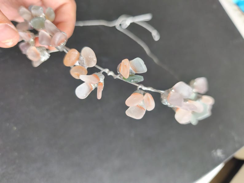 May include: A delicate floral hair accessory featuring small, translucent, pastel-colored stones. The stones are in shades of peach, pink, and green, attached to a silver-colored wire, creating a natural, organic look. The accessory is held by a hand.