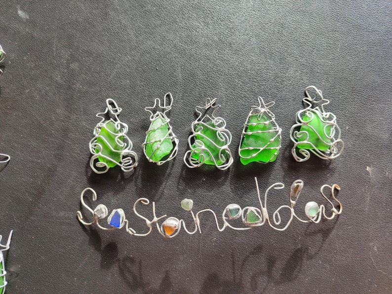 May include: Five green sea glass Christmas tree ornaments wrapped in silver wire. The word "Seaglass" is spelled out in silver wire below the ornaments.
