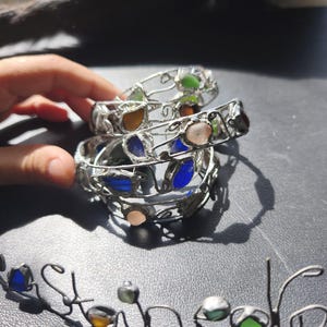 May include: A stack of silver-toned metal bracelets with colorful glass stones. The stones are in shades of blue, green, orange, and pink. The word "Stone" is spelled out in a matching silver-toned metal wire.