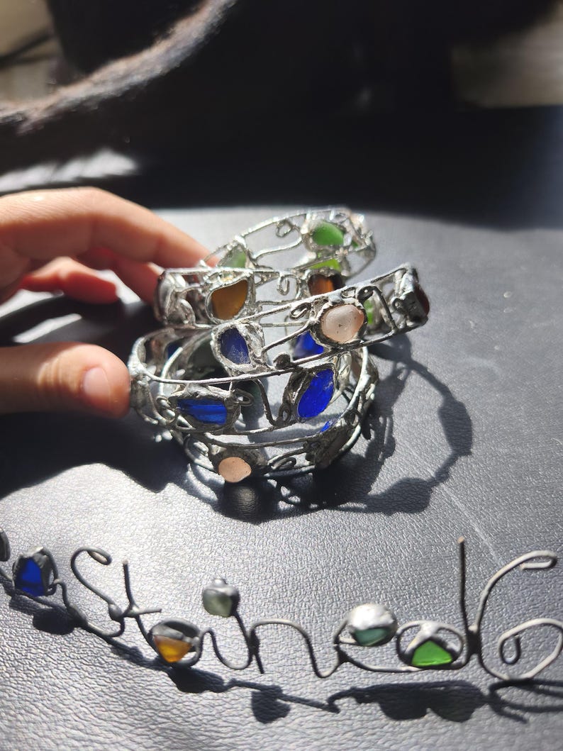 May include: A stack of silver-toned metal bracelets with colorful glass stones. The stones are in shades of blue, green, orange, and pink. The word "Stone" is spelled out in a matching silver-toned metal wire.