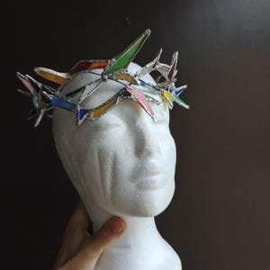 May include: A crown constructed from multi-colored stained glass shards held together by silver-toned metal solder. The crown features pointed, angular glass pieces in shades of green, blue, yellow, red, and pink, arranged in a jagged, circular formation.