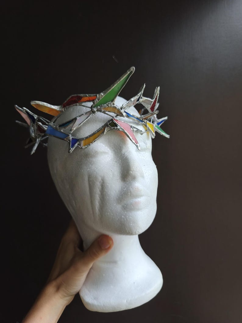 May include: A crown constructed from multi-colored stained glass shards held together by silver-toned metal solder. The crown features pointed, angular glass pieces in shades of green, blue, yellow, red, and pink, arranged in a jagged, circular formation.