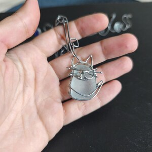 May include: A silver-toned cat pendant with a sea glass body and wire detailing. The cat has pointed ears, whiskers, and a curved tail. The pendant is suspended from a silver chain.