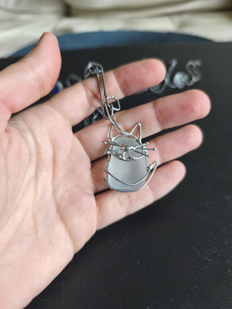 May include: A silver-toned cat pendant with a sea glass body and wire detailing. The cat has pointed ears, whiskers, and a curved tail. The pendant is suspended from a silver chain.