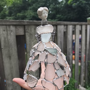 以下が含まれることがあります： 白い陶器の頭部と胴体を持ち、ワイヤーフレームのスカートに淡いピンクと透明なシーグラスの破片を銀色の金属ハンダでモザイク状に繋ぎ合わせた装飾用のフィギュア。シーグラスは半透明で、スカート全体に配置されている。